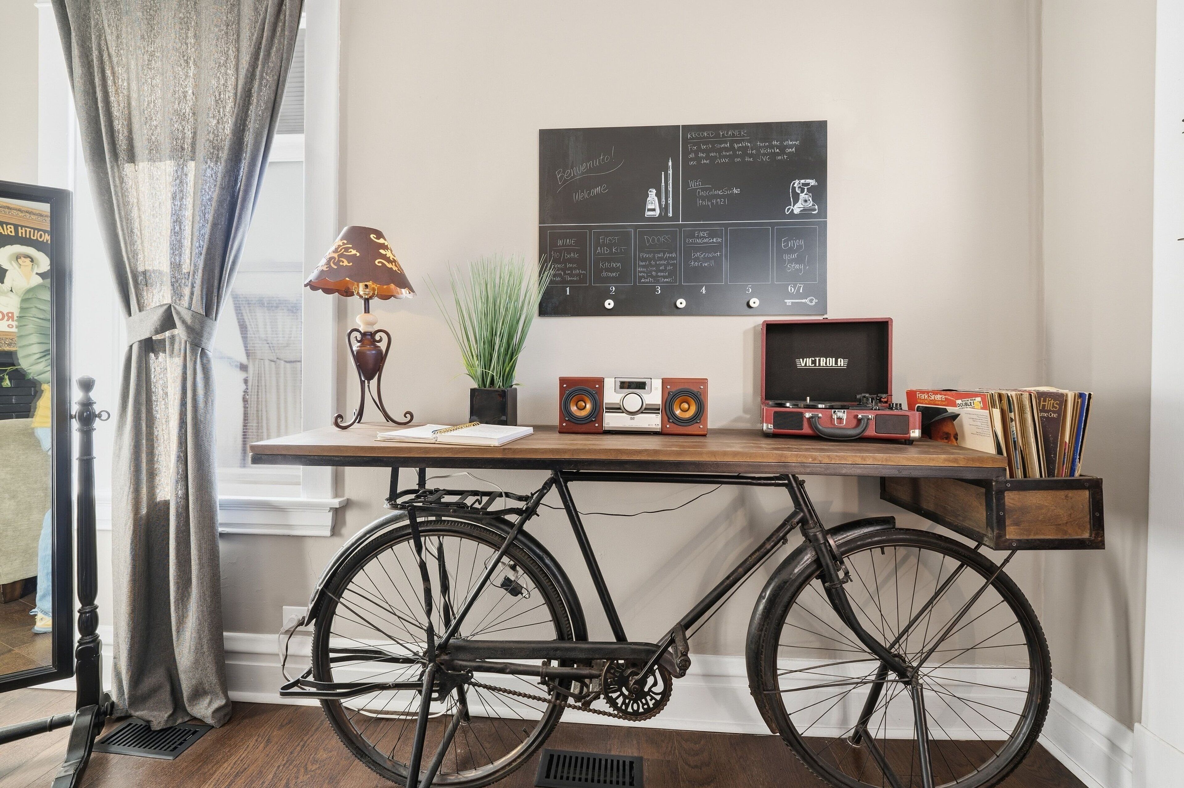 Una habitación con un escritorio de madera, una bicicleta antigua, una pizarra con un mensaje de bienvenida y una radio antigua.
