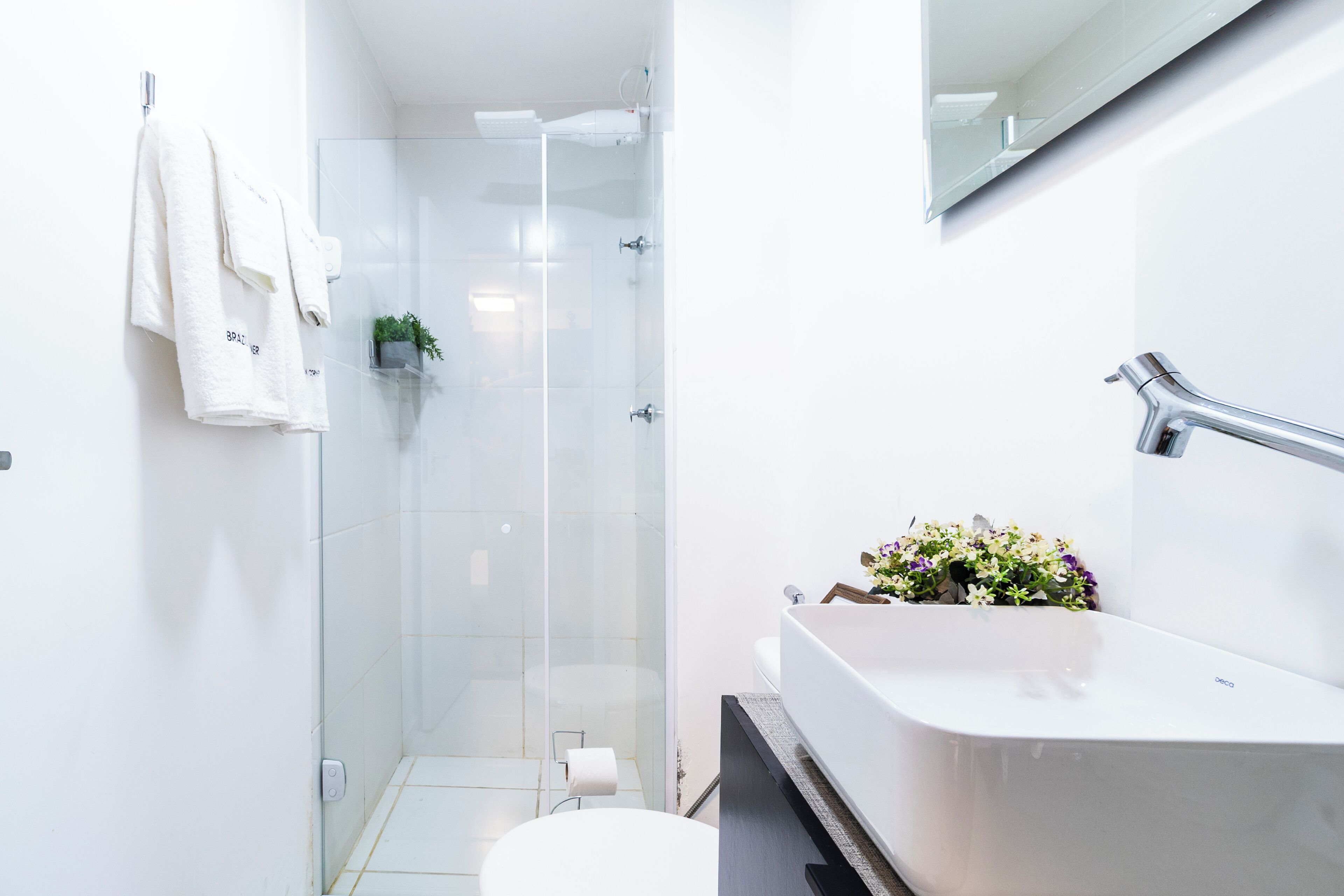 Un bany modern amb una cabina de dutxa de vidre, un lavabo blanc amb un arranjament floral i un para-tovalloles amb dues tovalloles.