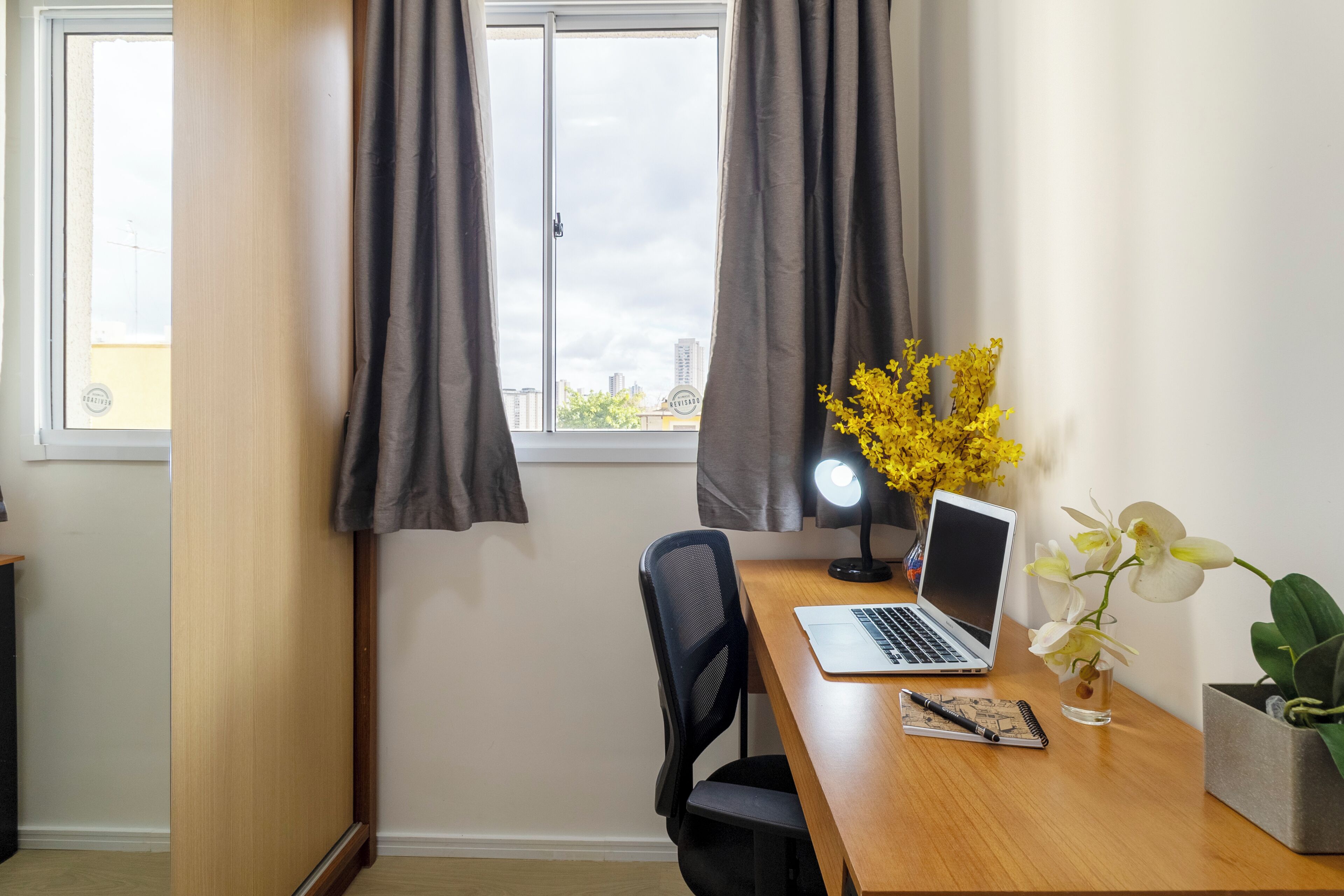 Ein Zimmer mit Schreibtisch aus Holz, einem Laptop, einem Notizbuch, einem Stift, einer Lampe und einer Vase mit Blumen.