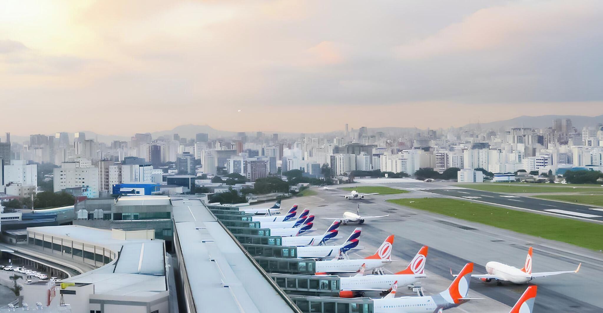 An airport terminal with multiple airplanes parked in the terminal area, a cityscape in the background, and a clear sky.