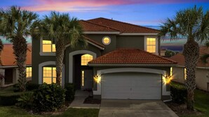 A two-story house with a red-tiled roof, a garage, and a well-maintained lawn with palm trees.