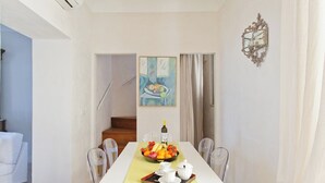 Un comedor con una mesa blanca dispuesta para dos personas, un frutero y una botella de vino.