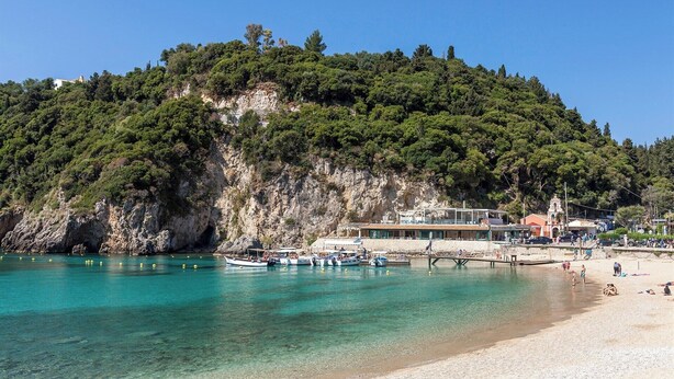Una zona costiera con spiaggia, acque turchesi cristalline, scogliere rocciose e un edificio con tetto rosso vicino alla riva.