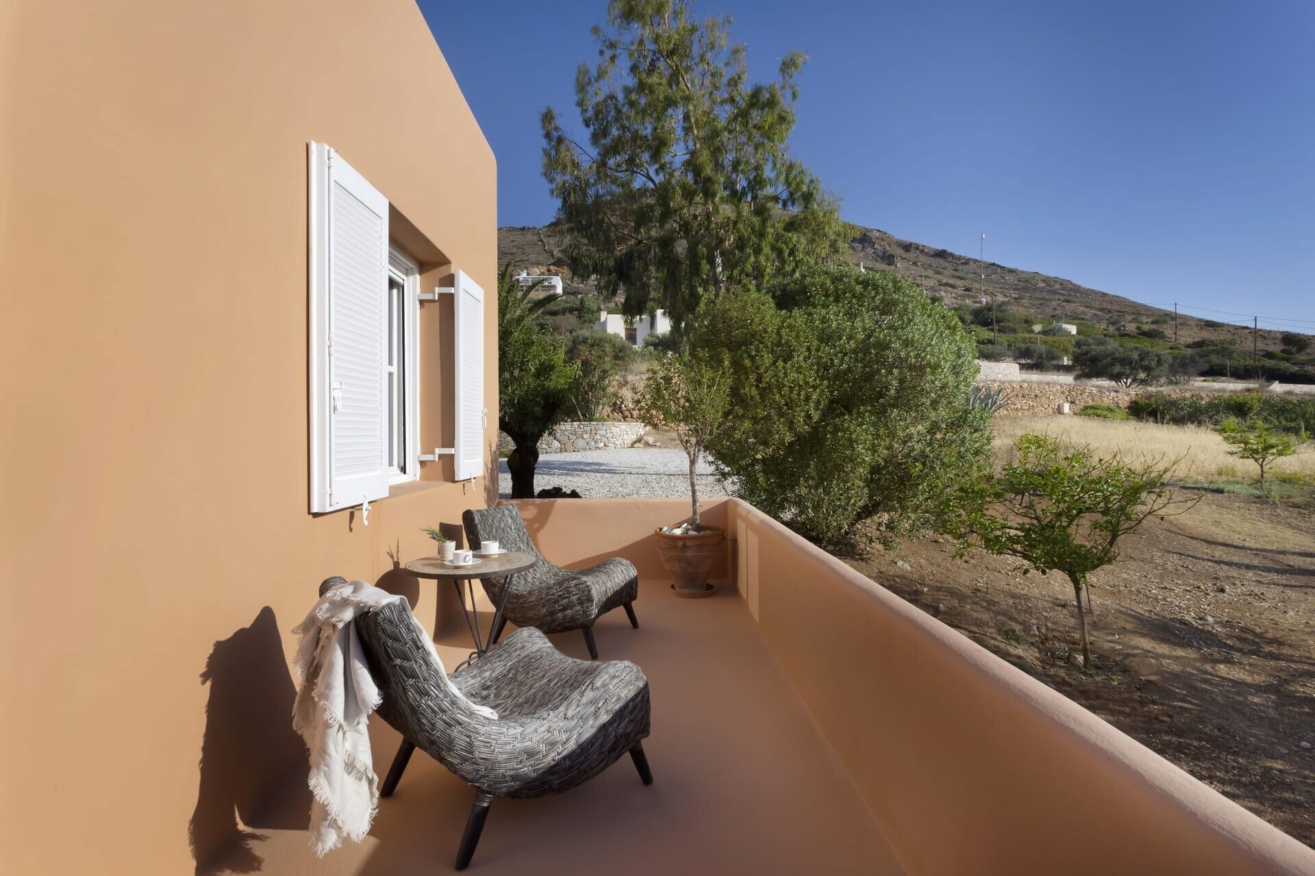 A balcony with two wicker chairs and a small table, overlooking a hillside with sparse vegetation and a clear sky.