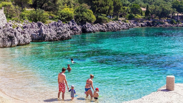 Una playa con agua turquesa cristalina, costa rocosa y abundante vegetación al fondo.