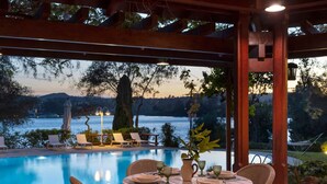 Un área de comedor junto a la piscina, con sillas de mimbre y una mesa preparada para dos, con vistas a un lago y montañas a lo lejos.
