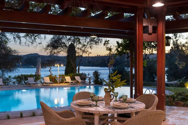A poolside dining area with wicker chairs and a table set for two, overlooking a lake with a view of the mountains in the distance.