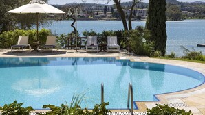 Una piscina con sillones de descanso y una sombrilla, rodeada de árboles y con vista a un lago y edificios al fondo.