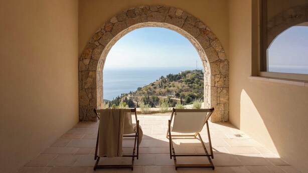 Un balcon agrémenté d’une arche en pierre, de deux chaises et offrant une vue sur un paysage côtier.