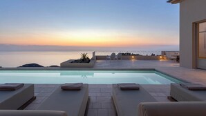 Un espace aménagé au bord de la piscine, avec des chaises longues, un petit coin salon et une vue sur la mer au coucher du soleil.