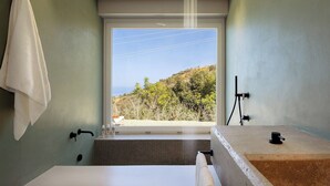 Un baño con bañera, ducha y lavamanos. Hay una toalla colgada en la pared, y una ventana con vista a árboles y colinas.