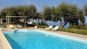 Piscina con sillones de descanso y una pérgola, rodeada de árboles y vistas al mar.