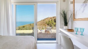 Un dormitorio con puerta corrediza de vidrio que da a un balcón con vista al mar y las colinas.
