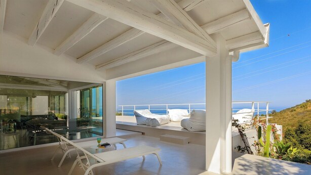 Interior moderno con techo blanco y pared de vidrio que da a un balcón con sillones blancos y vista al mar.