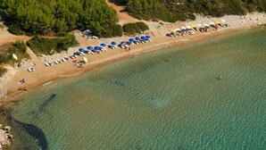 An aerial view of a beach with rows of umbrellas and sun loungers, clear shallow waters, and a rocky shoreline with vegetation.