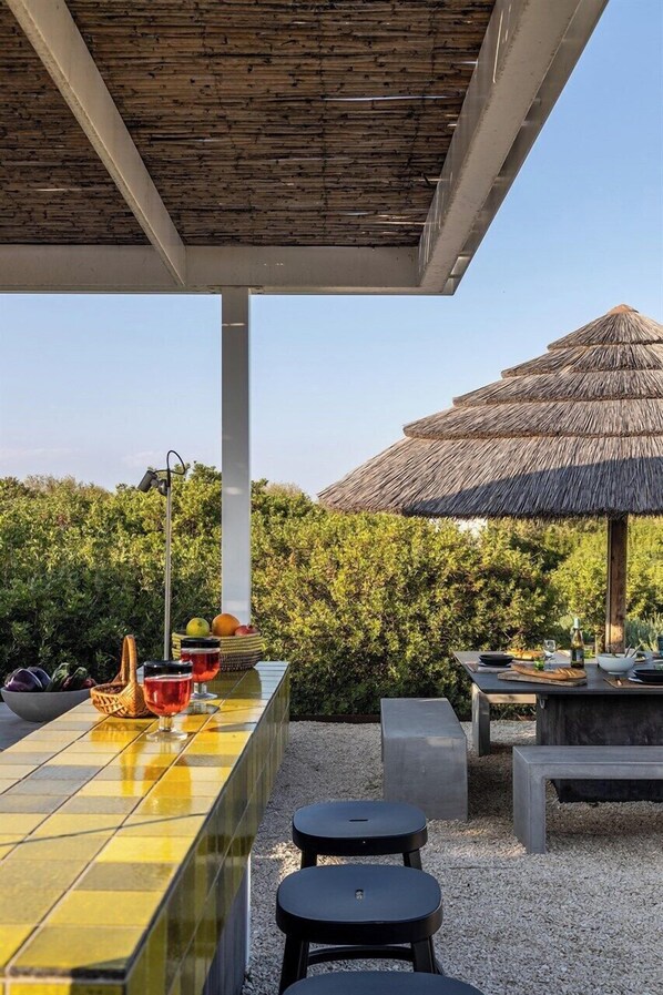 An outdoor dining area with a tiled bar, stools, and a thatched-roof structure.