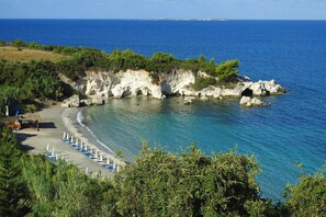 A coastal beach with clear blue waters, rocky cliffs, and a row of blue and white beach umbrellas.