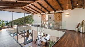 A modern interior with a glass floor leading to a balcony, wooden ceiling beams, and a view of a hillside.