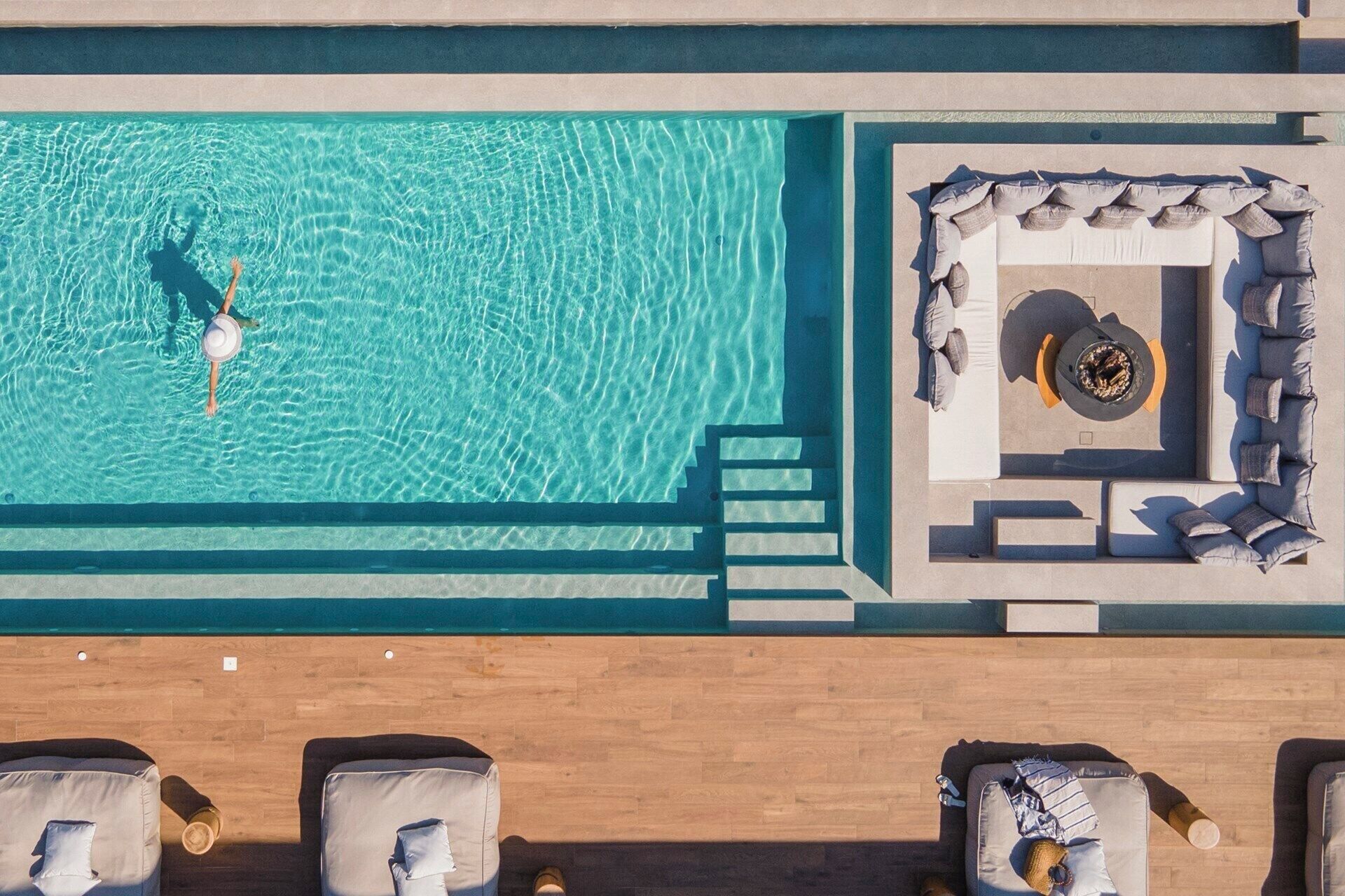 Vue aérienne d’un espace piscine avec une eau cristalline, un coin avec des chaises longues et un espace détente doté de coussins et d’un foyer.