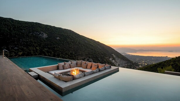 A infinity pool with a built-in seating area and a fire feature, overlooking a mountainous landscape during sunset.