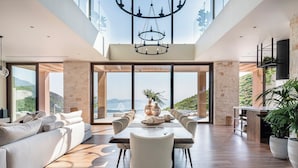 A spacious living area with a high ceiling, a dining table set for six, and a large window offering a view of the landscape.