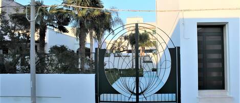 A gated entrance with a circular metal gate, a white building with a dark door, and palm trees in the background.