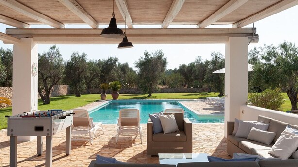 Uma área de piscina ao ar livre com mesa de pebolim, cadeiras de lounge e um sofá sob uma pérgola de madeira.