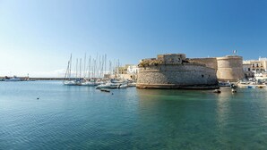 Uma marina costeira com uma fortaleza de pedra histórica, barcos e águas azul-claras.