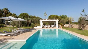 Piscina com espreguiçadeiras e guarda-sóis, cercada por vegetação e uma estrutura de pérgola.