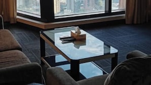 Ein Hotelzimmer mit großem Fenster und Blick auf eine Stadtlandschaft, darunter ein markanter Wolkenkratzer. Ein Glastisch mit Taschentuchbox und zwei Polsterstühle sind vorhanden.