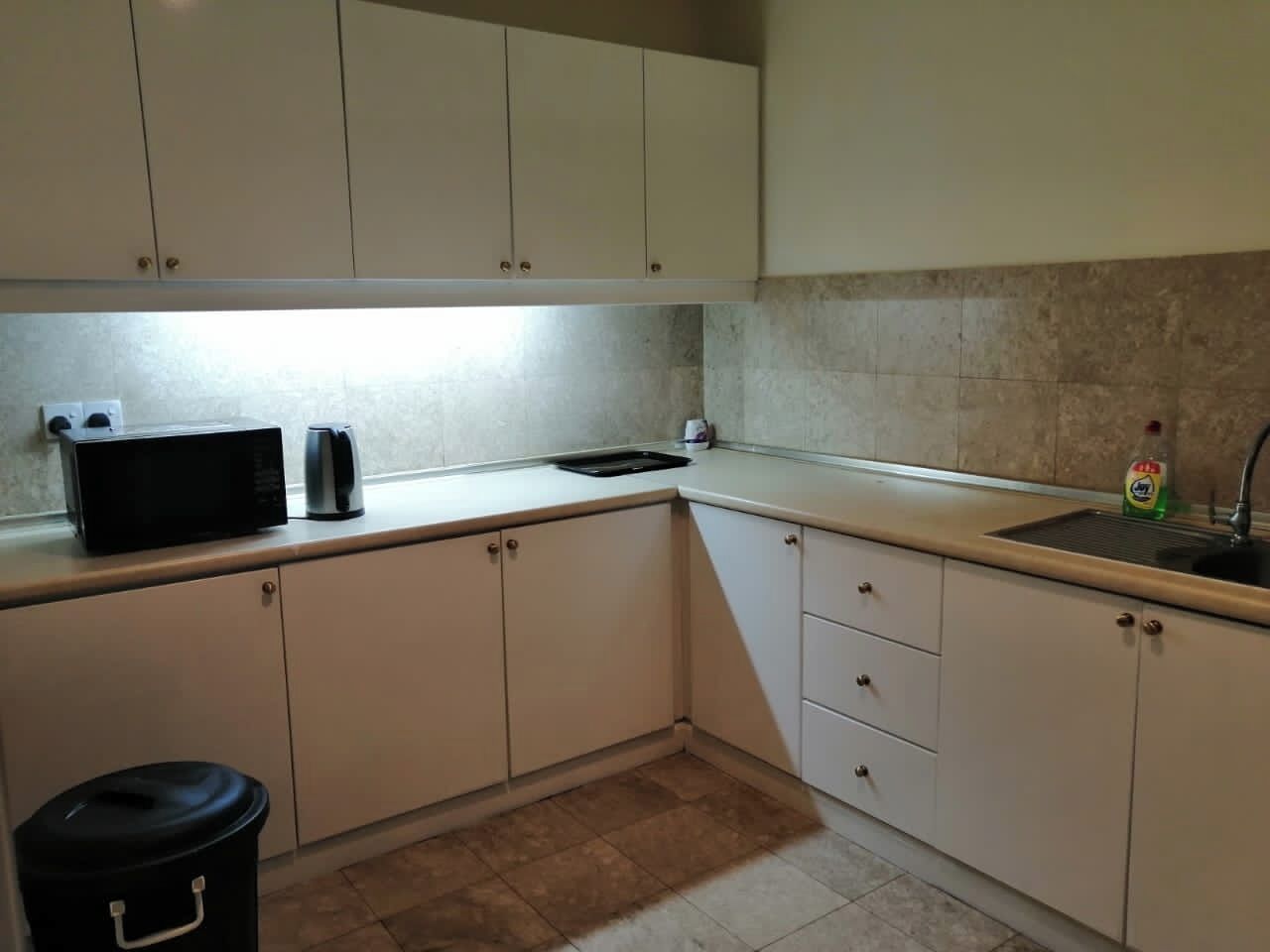 A kitchen with white cabinets, a black trash bin, a coffee maker, and a dish soap bottle.