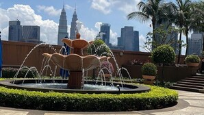 Ein Springbrunnen mit Wasserstrahlen in einer begrünten Anlage, mit Palmen und einer Stadtkulisse im Hintergrund.