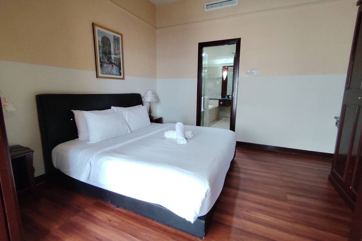 Una habitación de hotel con una cama grande, una mesita de noche, una lámpara, un cuadro en la pared y una puerta que da a un baño.