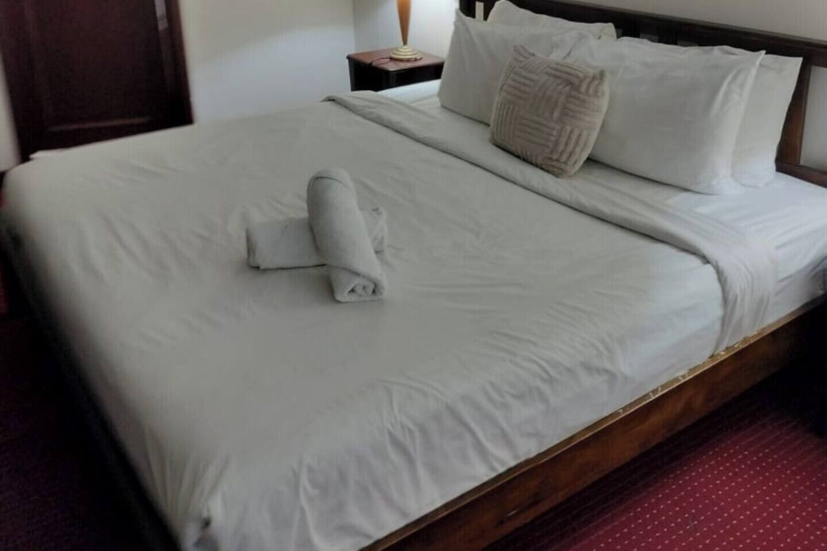 Una cama bien hecha con sábanas blancas, cabecera de madera, mesita de noche con lámpara y un cuadro floral en la pared.