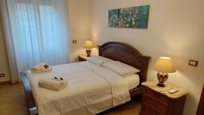 A neatly made bed with white linens and towels, two bedside lamps, a wooden nightstand, and a painting on the wall. - Alcinque - Your home in Rome, in the heart of San Giovanni (Roma)