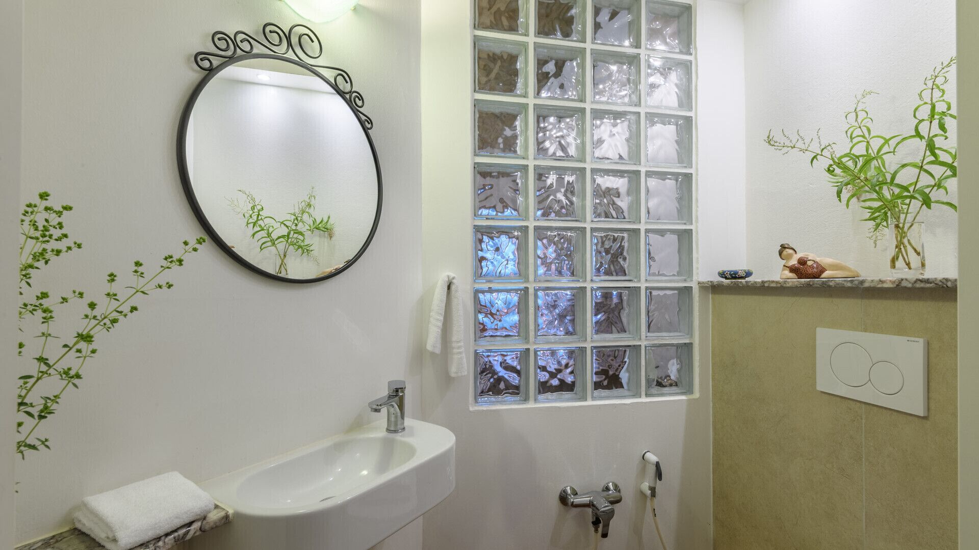 A bathroom with a round mirror, a sink, a shower, and a window with frosted glass panels.