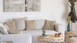 A modern living room with a sofa, coffee table, and chairs. There is a painting on the wall and a vase with plants on a side table.