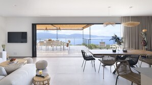 A modern living room with a large sliding glass door leading to a balcony with a view of the sea and mountains.