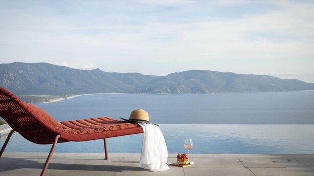 A lounge chair with a woven cushion, a straw hat, a white cloth, and a glass of wine on a terrace overlooking a calm sea and distant mountains.