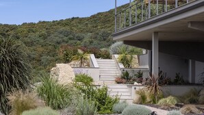A modern house with a stone staircase leading to a balcony, surrounded by various plants and a hillside in the background.