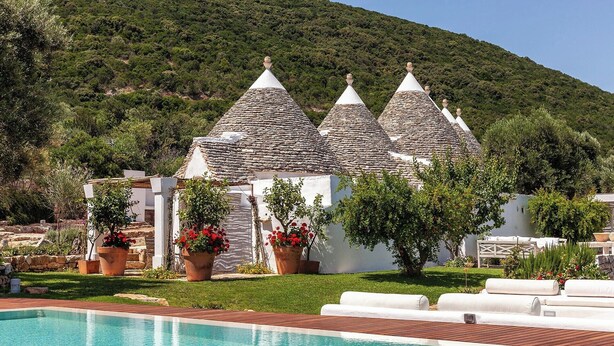A poolside area with a clear blue pool, white buildings with conical roofs, and lush greenery in the background. - I Trulli delle Sette Sorelle (Monopoli)