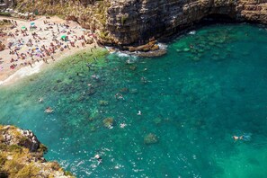 Ein Luftbild eines Strandes mit klarem türkisblauem Wasser, steinigen Klippen und einem Sandstrand, an dem Menschen schwimmen und sonnenbaden.