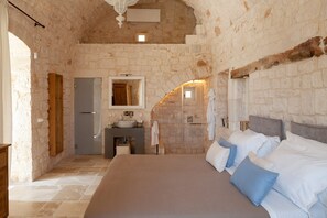 Ein Schlafzimmer mit Steinsandsteinwand, einem Bett mit weißblauen Kissen, einem Holzkopfende und einem Kamin aus Stein. Ein Badezimmer mit Waschbecken und Spiegel.