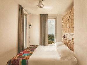 Un dormitorio con cama, cortinas, ventilador de techo, pared de piedra y vista al paisaje.