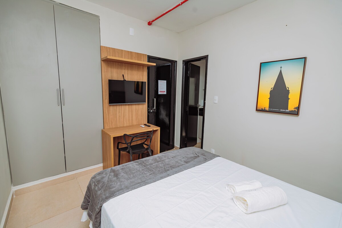 A bedroom with a bed, a desk, a chair, a TV, and a painting.