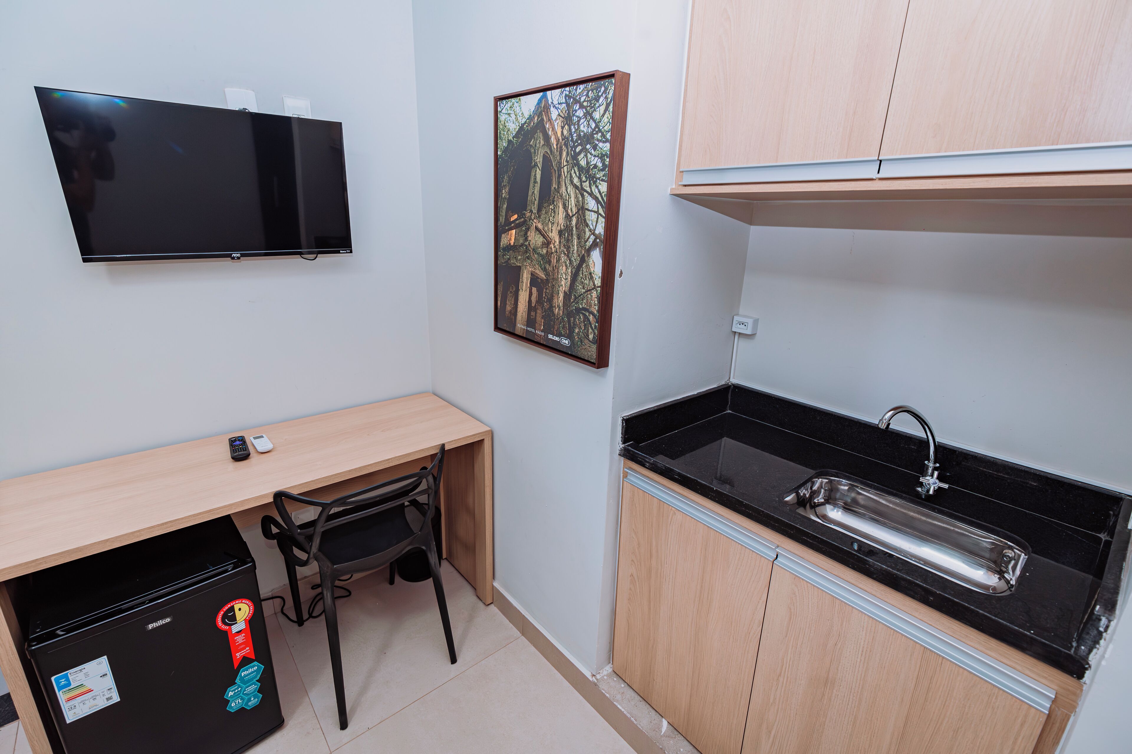 Uma kitchenette compacta com geladeira, pia, escrivaninha de madeira e televisão na parede.
