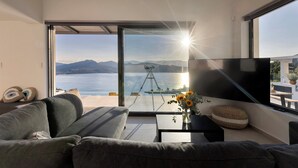 A modern living room with a large window overlooking a lake and mountains, a tripod with a camera, and a flat-screen TV. - Casa Rosemary - Two Bedroom Villa, Sleeps 4 (Lefkada City)
