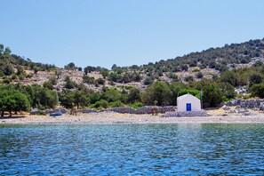 Un pequeño edificio blanco con puerta azul, rodeado de costa rocosa y árboles, con vista a una masa de agua tranquila.