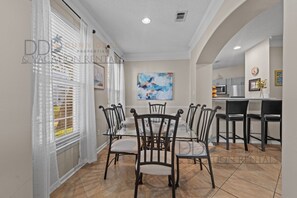 A dining area with a glass table and chairs, a painting on the wall, and a kitchen in the background.