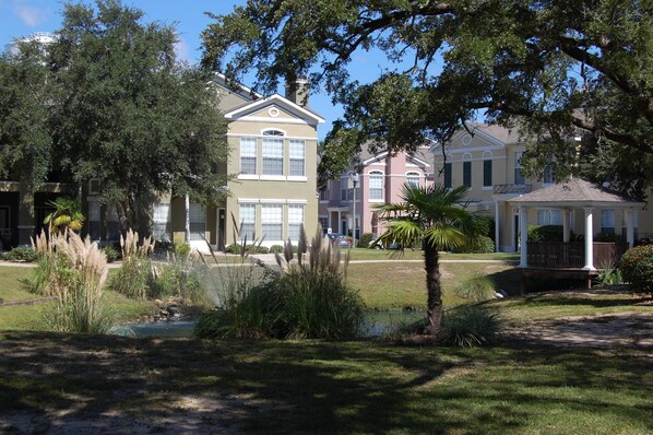A residential area with a large two-story house featuring a front porch, a small pond, and surrounding greenery.